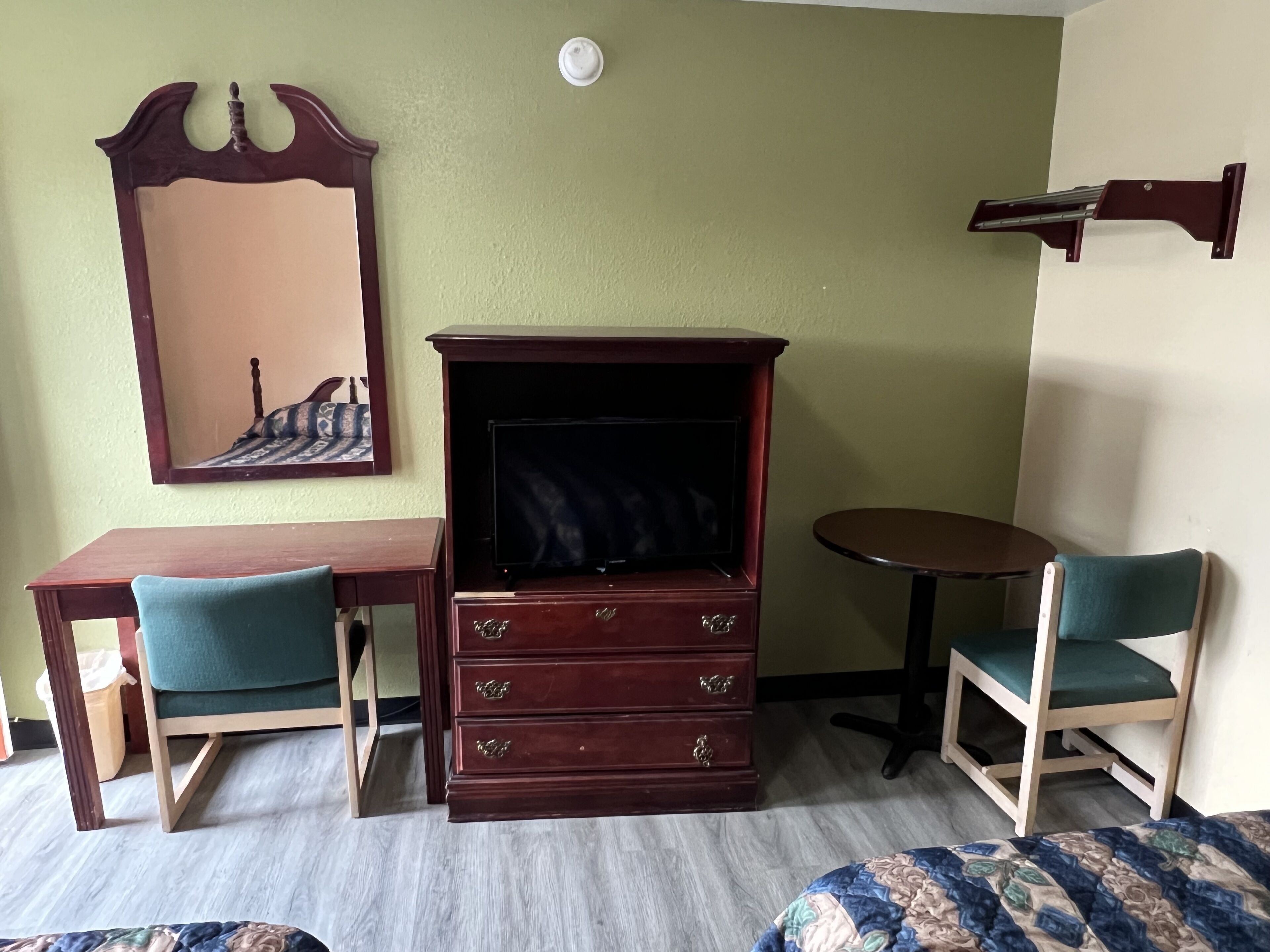 A room with a wooden desk, a dresser with drawers, a chair, a bed, and a wall-mounted shelf.