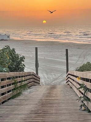 Ein hölzerner Steg führt zu einem Strand, im Hintergrund ein Sonnenuntergang.