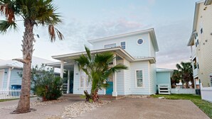 A two-story house with a front porch, palm trees, and a white picket fence. - Shared Pool and Hot Tub, Golf Cart Included, Beach Boardwalk, Pet Friendly (Port Aransas)