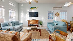 A spacious living room with two light blue sofas, a wooden coffee table, wicker chairs, a ceiling fan, and a mounted TV. - Shared Pool and Hot Tub, Golf Cart Included, Beach Boardwalk, Pet Friendly (Port Aransas)