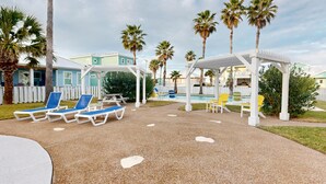 A poolside area with lounge chairs, a covered seating area, and palm trees. - Shared Pool and Hot Tub, Golf Cart Included, Beach Boardwalk, Pet Friendly (Port Aransas)