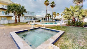 A swimming pool with lounge chairs, palm trees, and a two-story building with a white fence. - Shared Pool and Hot Tub, Golf Cart Included, Beach Boardwalk, Pet Friendly (Port Aransas)
