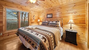 A wooden cabin bedroom with a large bed, a nightstand, a ceiling fan, and a window with a view of trees. - Cabin with Appalachian Mountain Views (Boone)