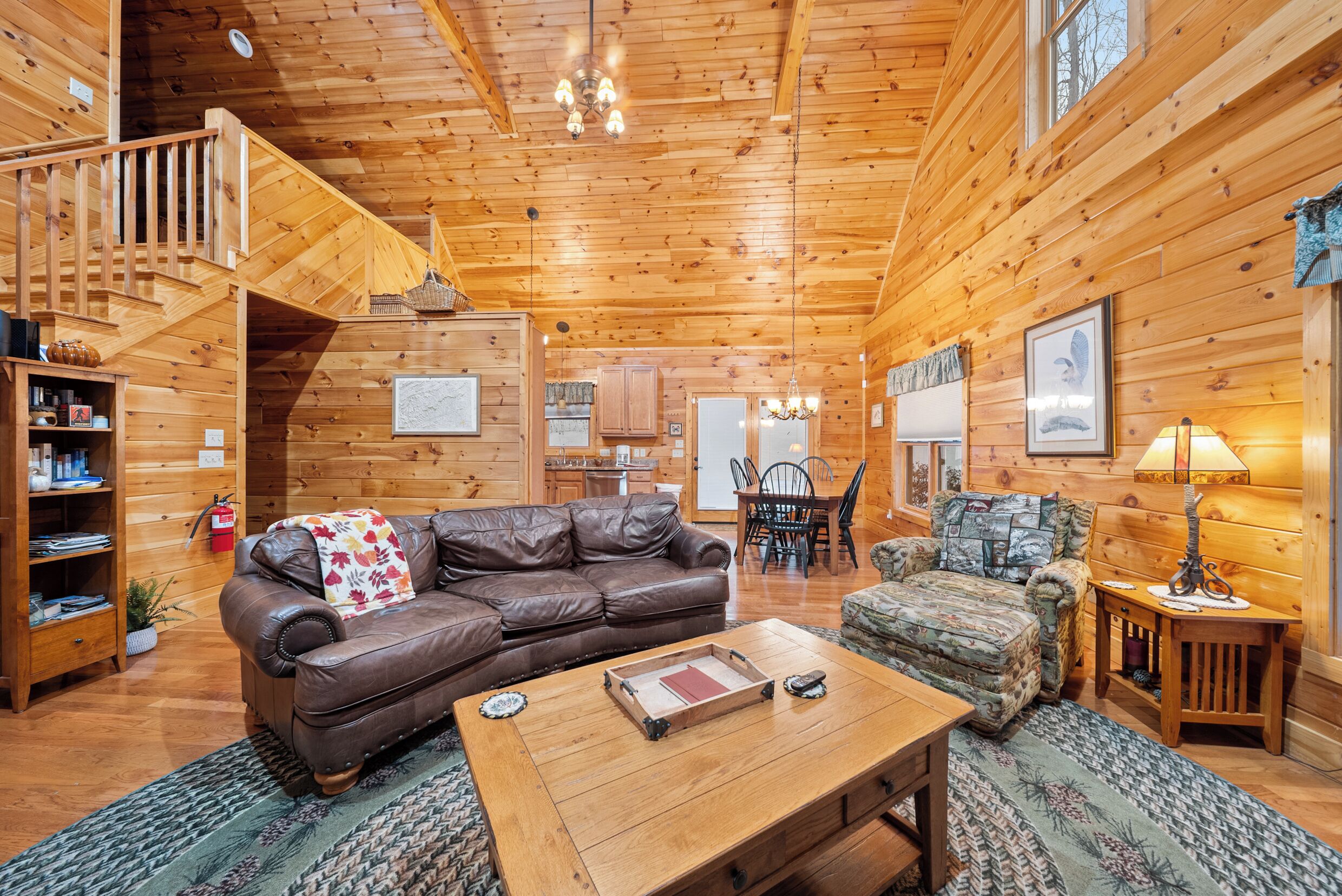 A cozy log cabin interior with a leather sofa, a wooden coffee table, a patterned armchair, and a dining area with a table and chairs.