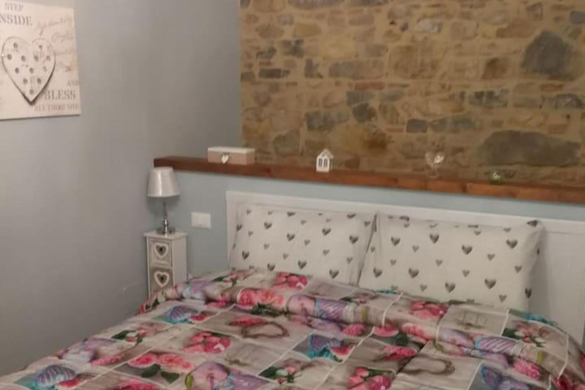 Un dormitori amb paret de pedra, un llit amb roba de llit floral, una tauleta de nit amb una làmpada i una decoració amb forma de cor a la paret.