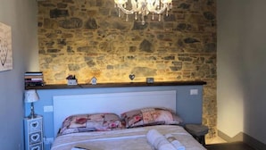 A bedroom with a stone wall, a chandelier, a bed with floral bedding, a bedside table with a lamp, and a small shelf with books and decorative items. - Beautiful Lux Apartment Chianti Firenze Area Italy (Barberino Tavarnelle)