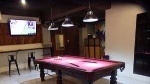 Une table de billard recouverte d’un feutre rose, deux lampes suspendues au plafond et un téléviseur fixé au mur.