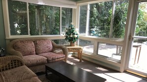 A cozy living room with a wicker sofa, a dark coffee table, and a small wooden side table with a plant. Large windows provide a view of the outdoors. - Enjoy Florida in Illinois; House on Island with Pool (East Moline)