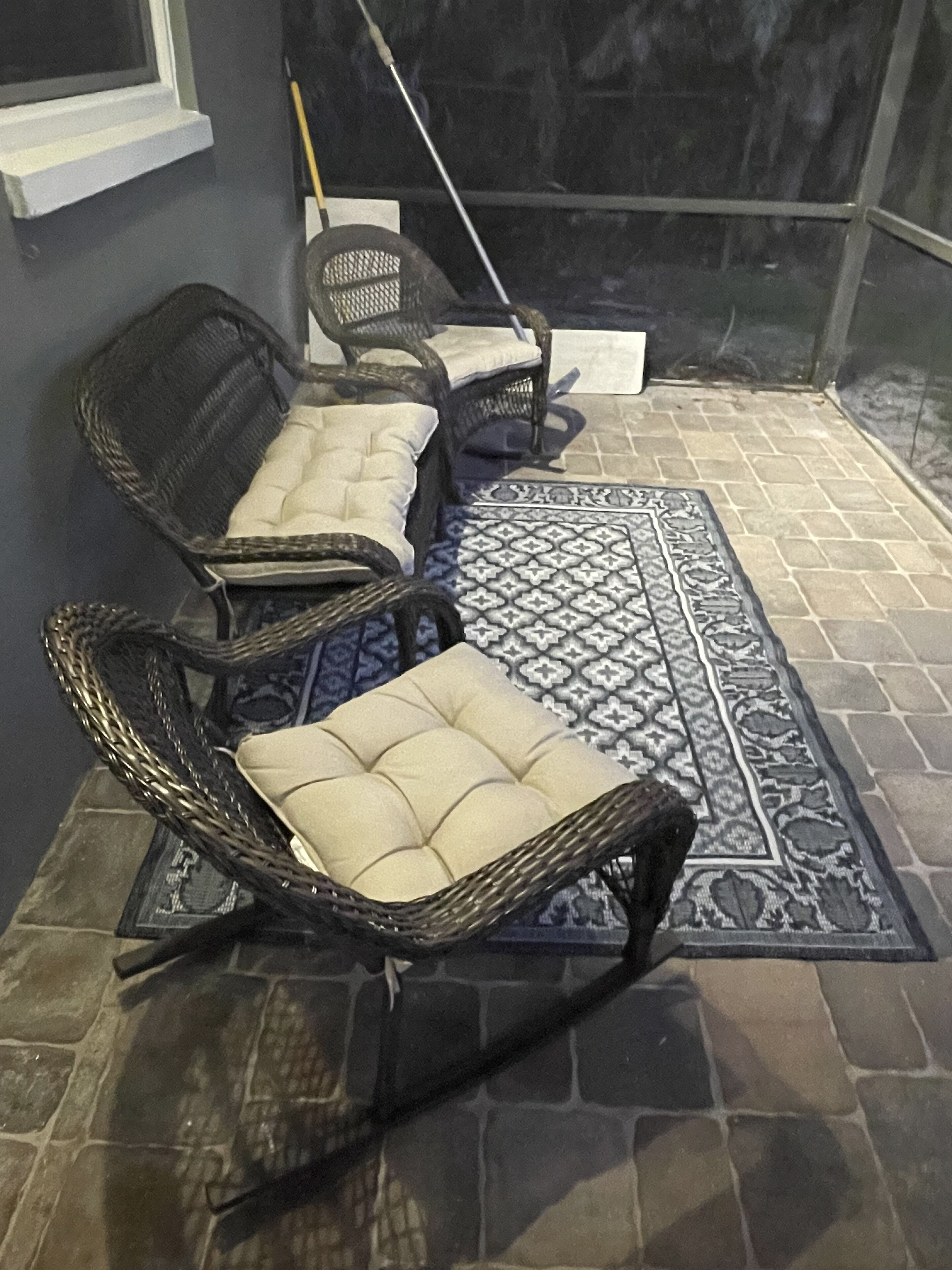 Wicker stoelen met kussens, een vloerkleed met patroon en een bezem die tegen een muur staat.