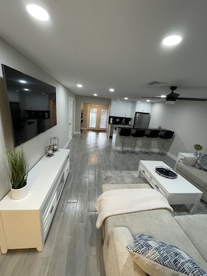 Una sala de estar moderna con una televisión de pantalla plana, un sofá, una mesa de centro y una zona de cocina con refrigerador y armarios.