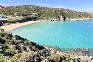 Paisatge costaner amb mar turquesa cristal·lina, terreny rocos i alguns edificis al fons.