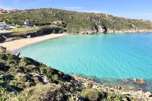 A coastal landscape with a clear turquoise sea, rocky terrain, and a few buildings in the background.