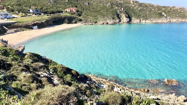 A coastal landscape with a clear turquoise sea, rocky terrain, and a few buildings in the background.