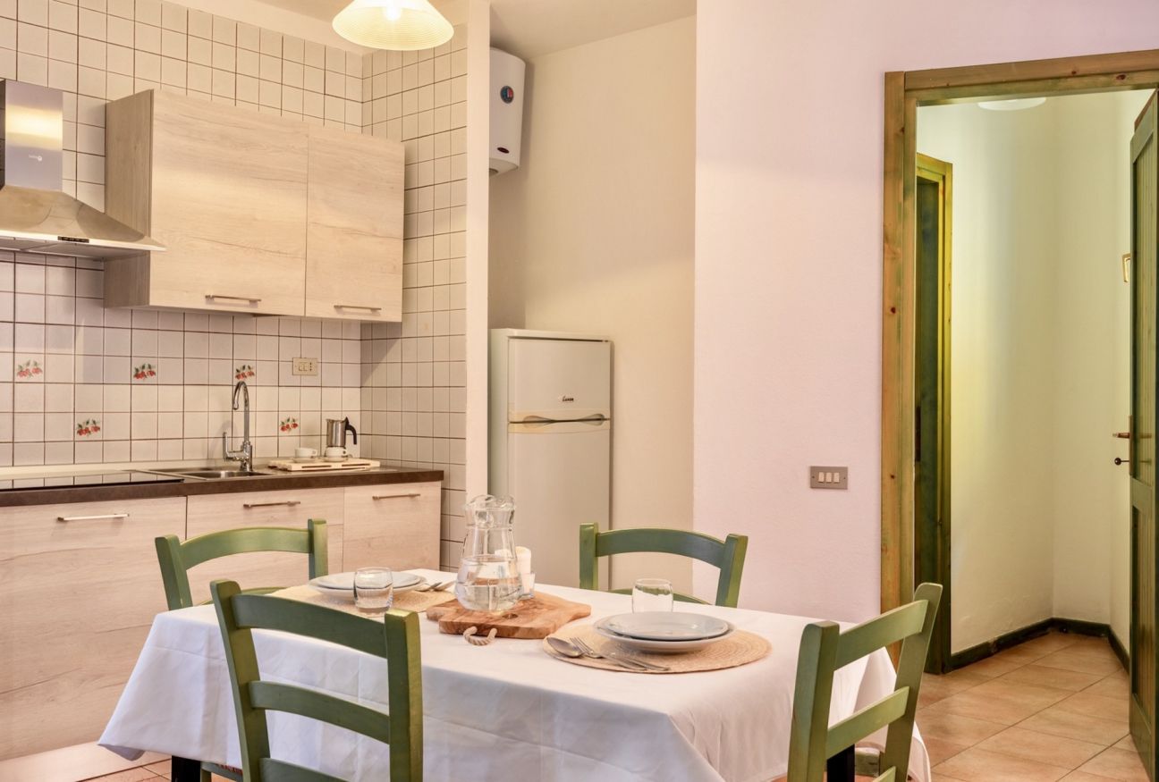 Un coin repas avec une table dressée pour deux personnes, une cuisine aux carreaux blancs et aux meubles en bois, et une porte donnant sur une autre pièce.