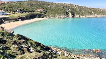 A coastal landscape with a clear turquoise sea, rocky terrain, and a few buildings in the background.