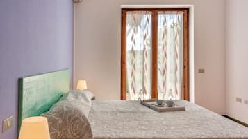 Un dormitorio con cama, lámpara de noche, bandeja con taza y platillo, y ventana con cortinas.