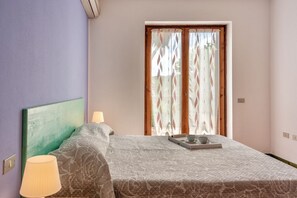 Un dormitorio con cama, lámpara de noche, bandeja con taza y platillo, y ventana con cortinas.