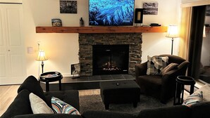 Un salon doté d’une cheminée en pierre, d’un téléviseur à écran plat fix é au-dessus, et d’un coin salon confortable avec un canapé et un pouf.