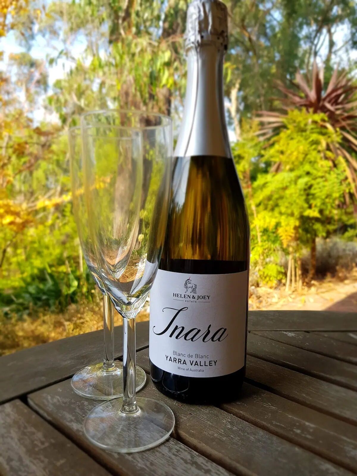 Una botella de vino Inara y dos copas sobre una mesa de madera con un fondo natural al aire libre.