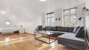 Moderni loft-tyylinen asunto, jossa on suuri harmaa kulmasohva, lasinen sohvapöytä ja valkoinen työpöytä tuolilla.