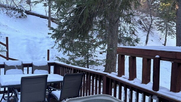 A hot tub with a wooden deck and chairs, surrounded by snow-covered trees.