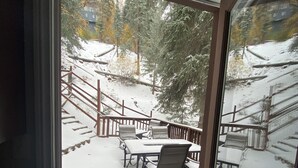 A snow-covered patio with outdoor furniture, a wooden deck, and a view of trees and a building in the background.