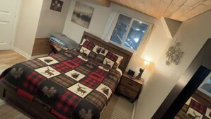 A bedroom with a wooden ceiling, a bed with a quilt, a nightstand, a window, and a wall-mounted light fixture.
