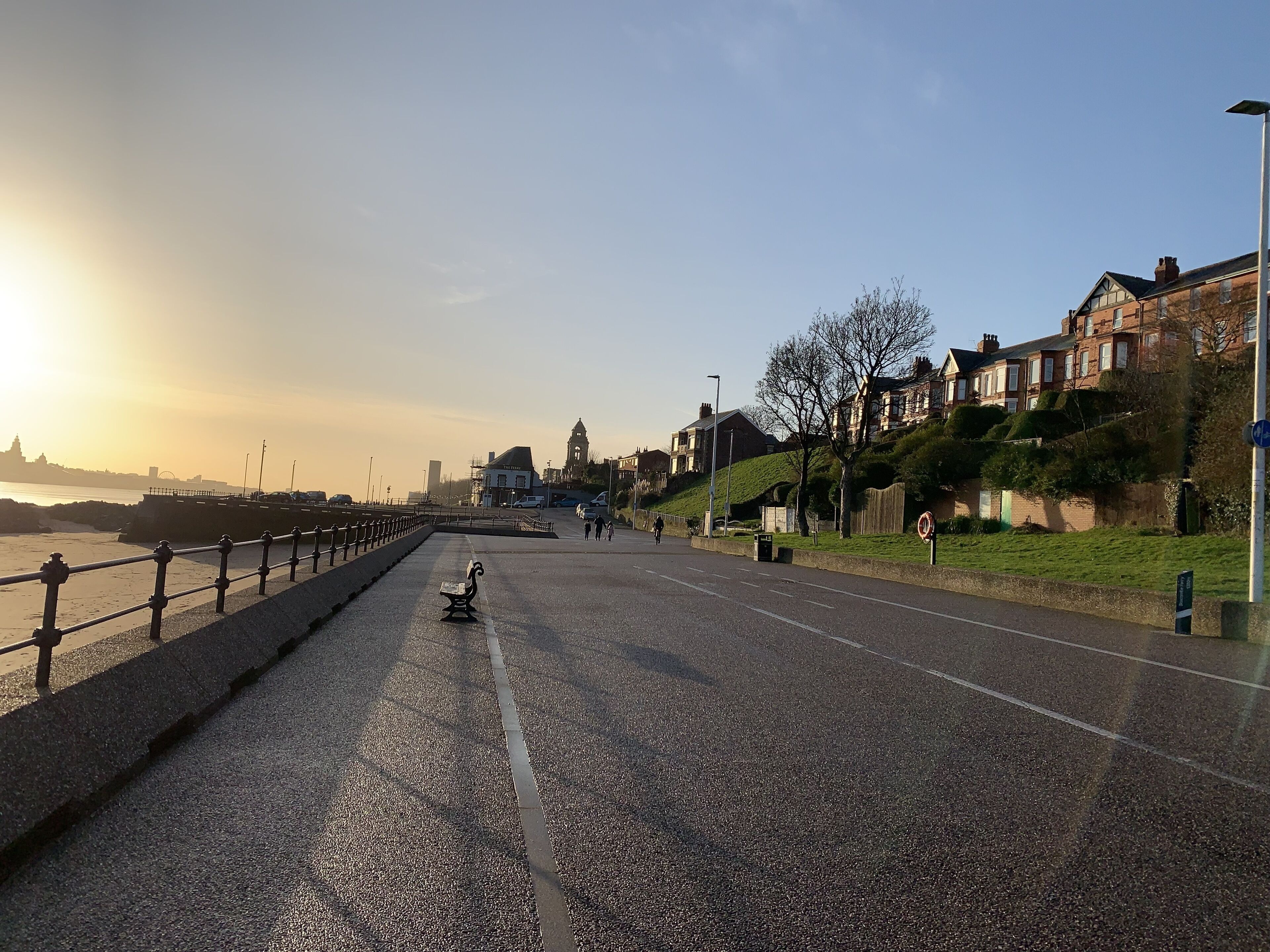 A bridge with a metal railing, a bench, and a road leading to a hill with houses.