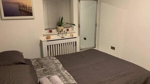 Un dormitorio con cama, una mesita de noche con una planta, un espejo y una ventana con persianas.
