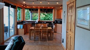 A cozy kitchen with wooden cabinets, a dining table, and chairs. There are large windows providing a view of the outdoors.