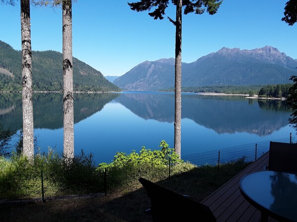 A tranquil lakeside view with a wooden deck and a table, surrounded by tall trees and mountains in the background. - Spectacular Views from every room
Right on the Lake - Now with Air Conditioning! (Hoodsport)