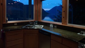 A kitchen with wooden cabinets, a stainless steel sink, and two windows offering views of a lake and mountains at dusk.