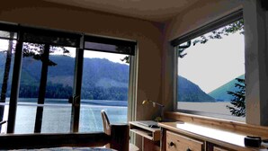 A room with a desk, chair, and large windows offering a view of a lake and mountains.