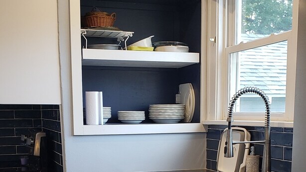 Una cocina compacta con armarios blancos, un estante empotrado con vajilla, un lavavajillas de acero inoxidable y una ventana con cortina.