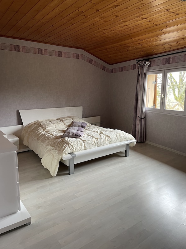 A bedroom with a wooden ceiling, a bed with a white frame, a bedside table, and a window with curtains.