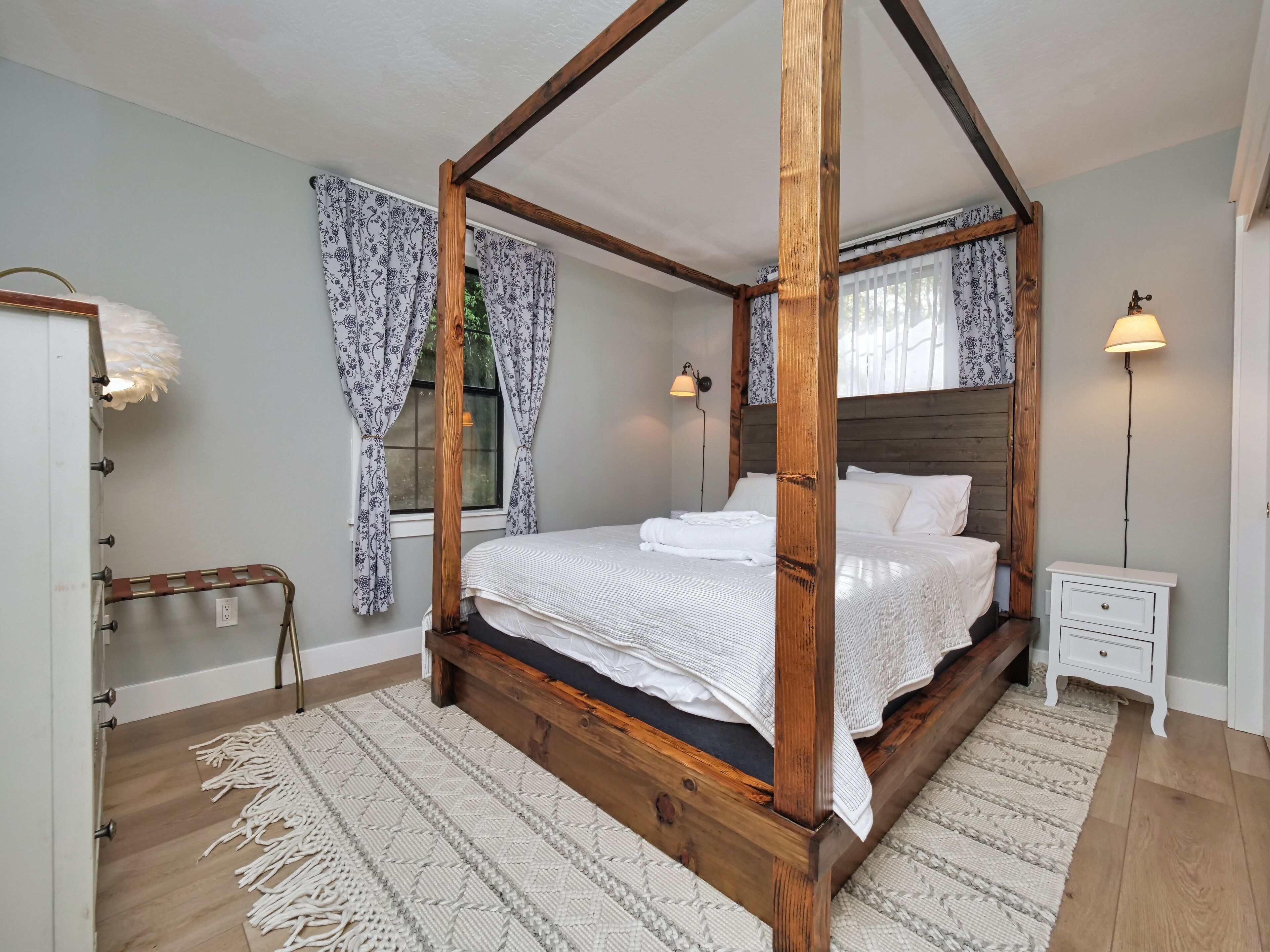 A four-poster bed with a wooden frame, white bedding, and a patterned rug. There are two bedside tables with lamps, a wooden bench, and a window with patterned curtains.