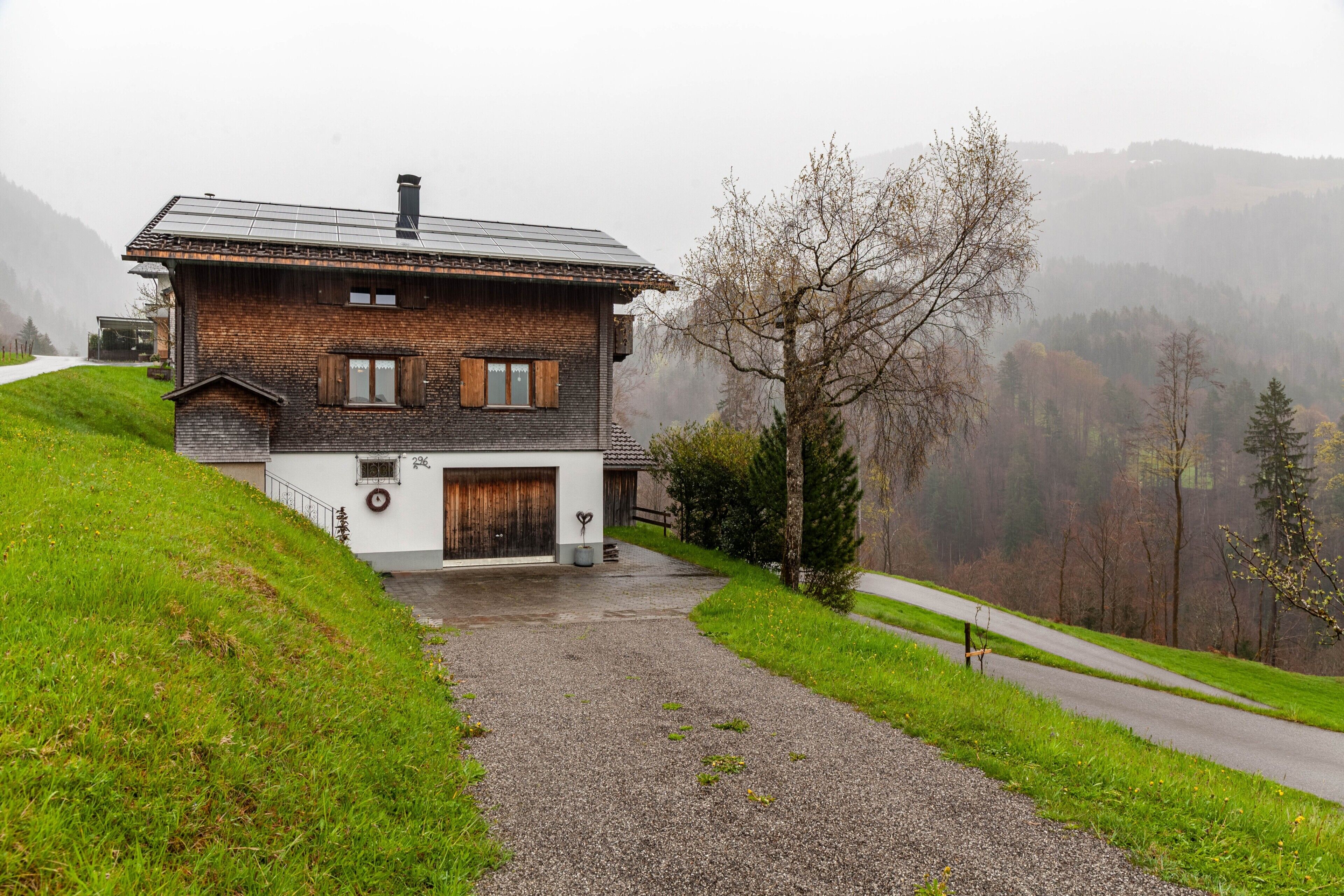 A two-story house with a sloped roof, a gravel driveway, and a view of a forested valley.