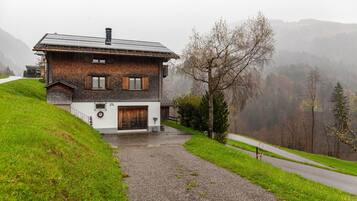 A two-story house with a sloped roof, a gravel driveway, and a view of a forested valley.