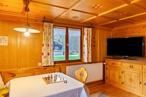 A wooden cabin room with a bed, a chessboard, a television, and a window with curtains. - Vacation Home "Dünser" with Mountain View, on a sunny slope. (Bizau)