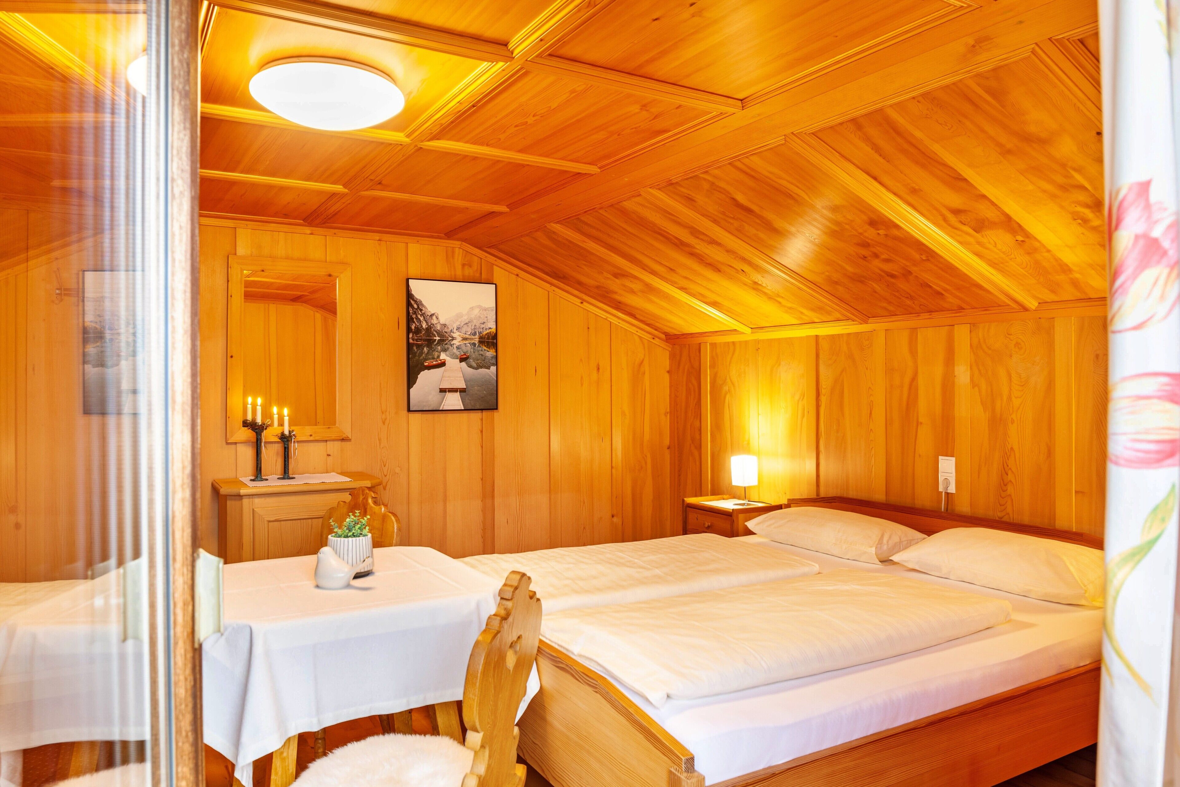 A wooden room with a bed, a table with a vase, and a painting on the wall.