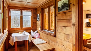A wooden cabin interior with a dining table, bench, and windows overlooking a snowy landscape. - Vacation Home "Dünser" with Mountain View, on a sunny slope. (Bizau)