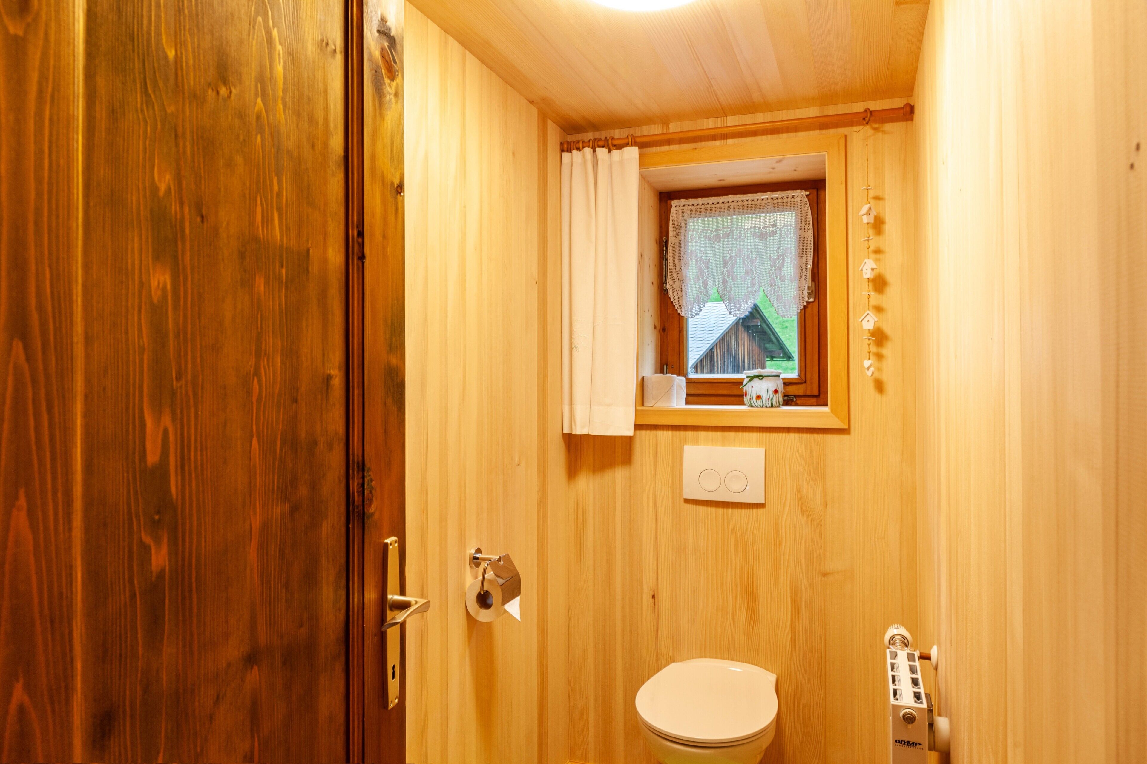 A wooden bathroom with a toilet, a window with a curtain, and a visible light switch.