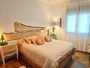 A bedroom with a wooden headboard, a bedside table with a lamp, a decorative wall piece, and a window with curtains. - Villa Zahara (Chiclana de la Frontera) (Chiclana)