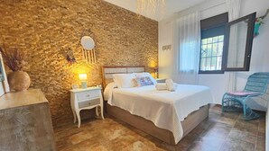 Une chambre avec un mur en pierre, un lit, une table de chevet, une chaise et une fenêtre.