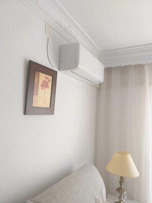 A hotel room with a bed, a bedside table with a lamp, a framed picture on the wall, and an air conditioning unit.