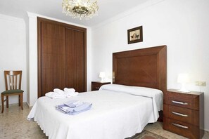 A hotel room with a large wooden wardrobe, a bed with white linens, a chair, and a bedside table with a lamp.