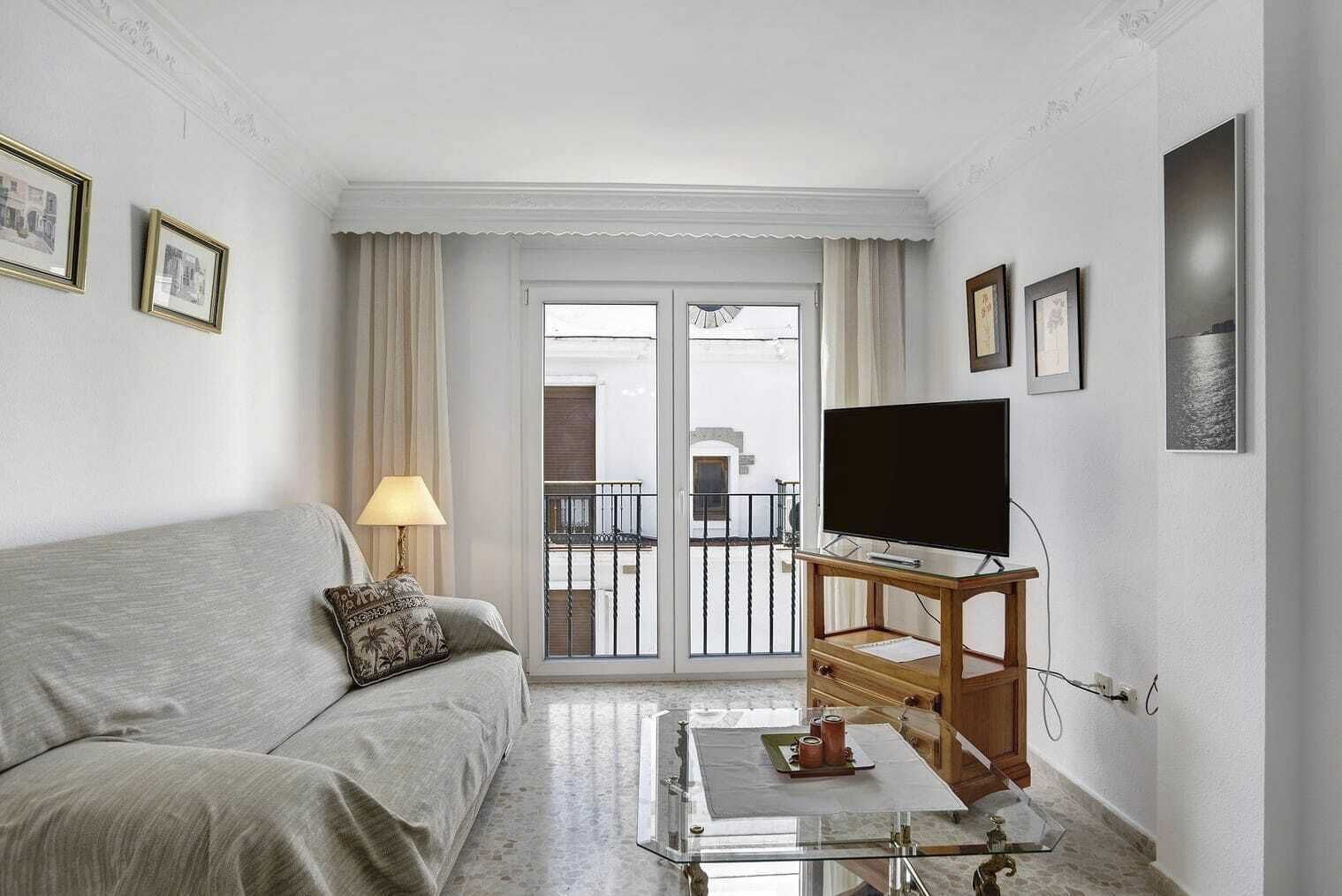 Un salon avec un canapé, un téléviseur posé sur un meuble en bois, une table basse en verre et un balcon doté d’une rambarde.