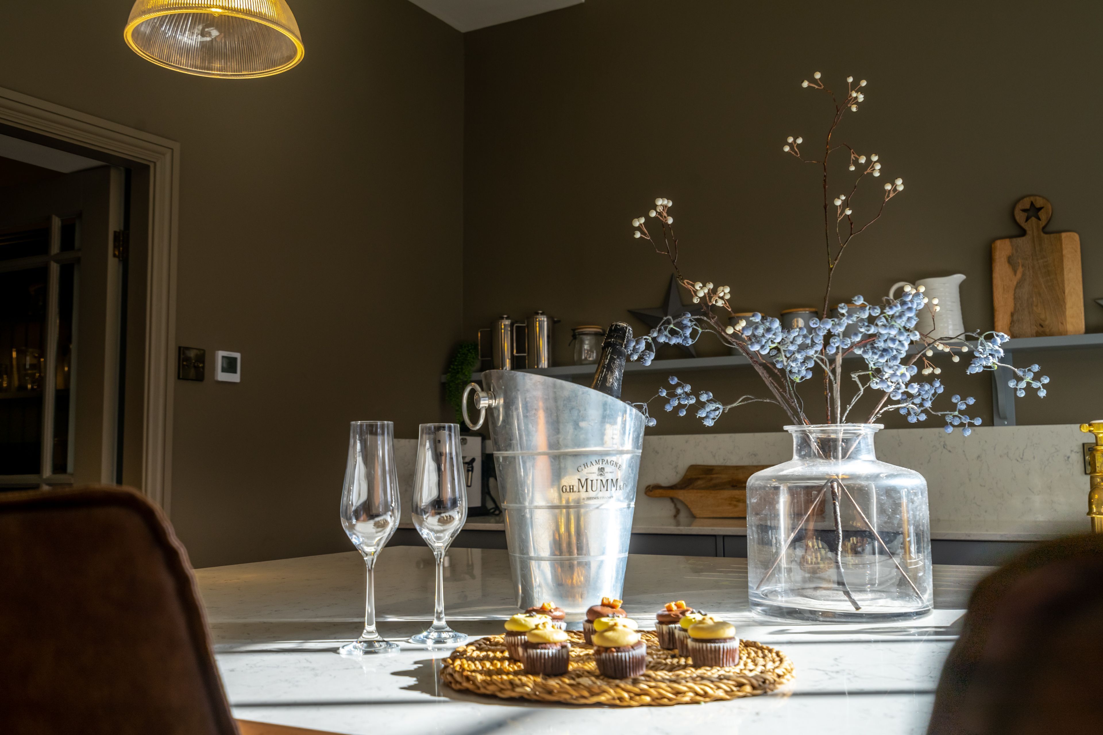 A dining table with a metal bucket, a glass vase with flowers, and a wooden cutting board.