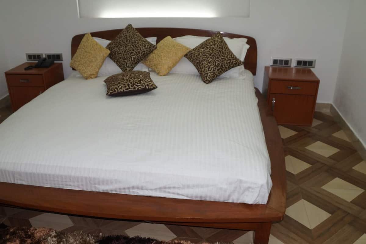 A neatly made bed with a white bedspread and patterned pillows, wooden headboard, two bedside tables with a phone and a lamp, and a patterned floor.