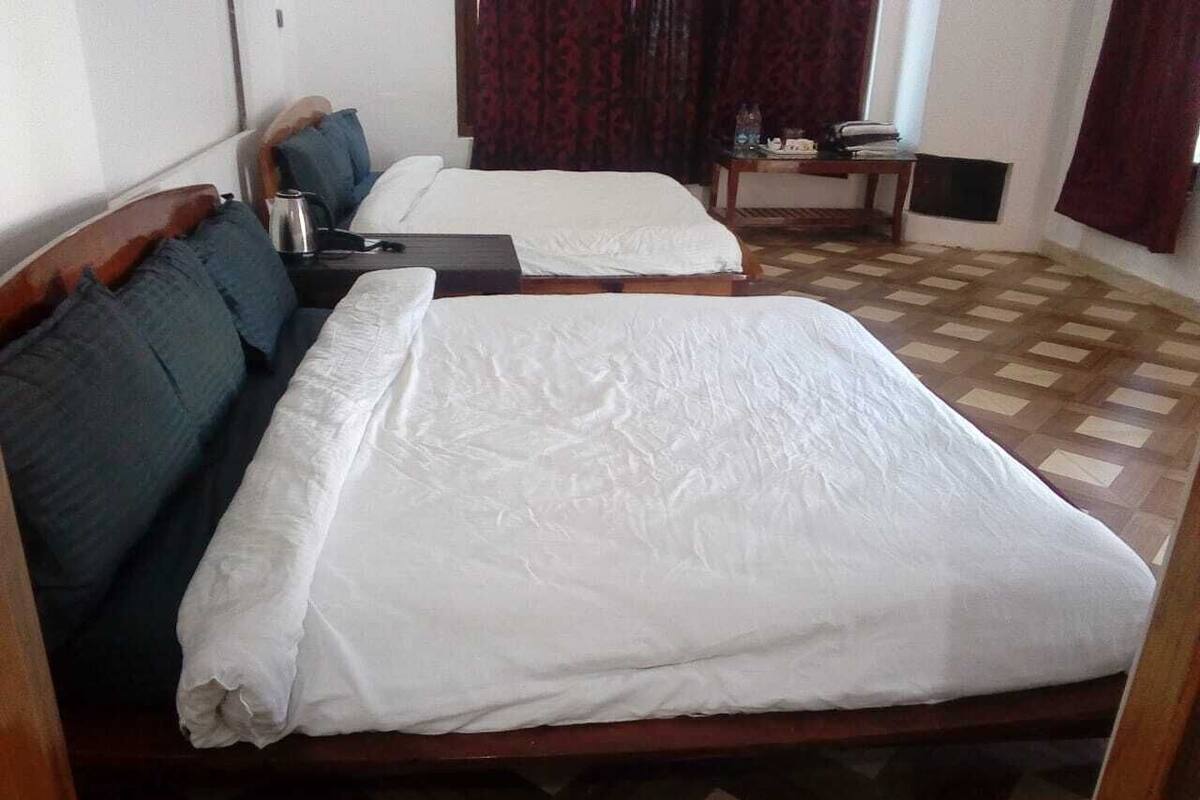 A room with two beds, each with white linens and wooden headboards. There is a small table with a lamp and a bottle of water. The room has a checkered floor and a window with curtains.